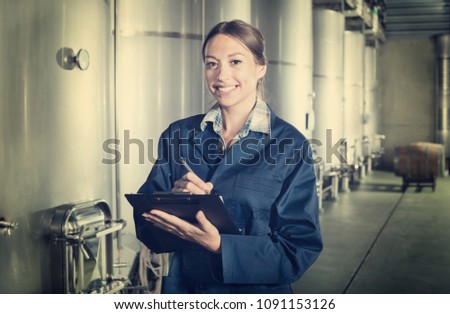 Young cheerful woman wearing a coat taking notes in secondary fermenting section on the wine factory