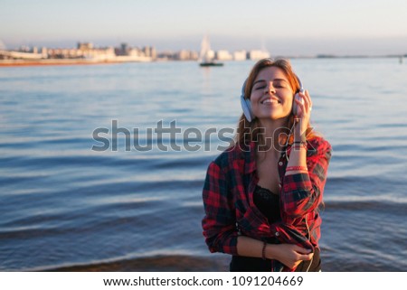 Happy attractive red-haired girl is enjoying favorite music on the walk. Beautiful young woman in shirt uses headphones
