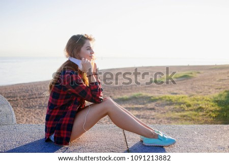Beautiful young redheaded woman sitting on the waterfront, enjoying the nature and the good weather. The girl in the red plaid shirt