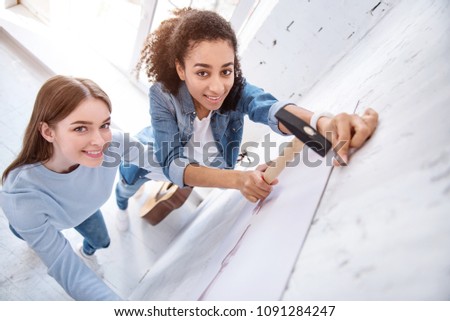 Art lovers. The top view of beautiful young girls smiling at the camera while hammering in a nail and hanging a picture on the wall