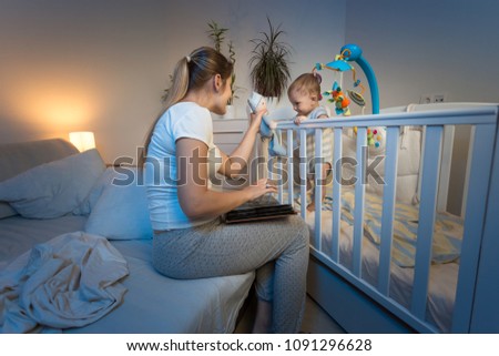 Young mother reading book aand giving toy to her baby son standing in cot at night