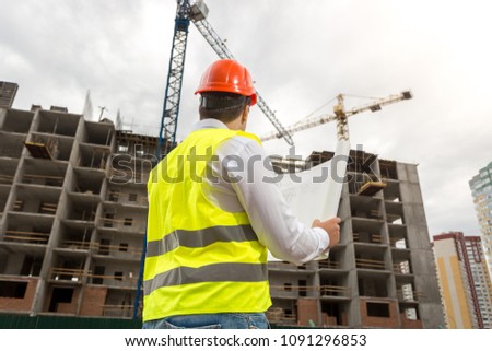 Rear view photo of male architect in hardhat and vest looking at working building cranes