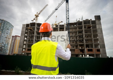 Rear view image of male construction engineer with blueprints controlling work on building site