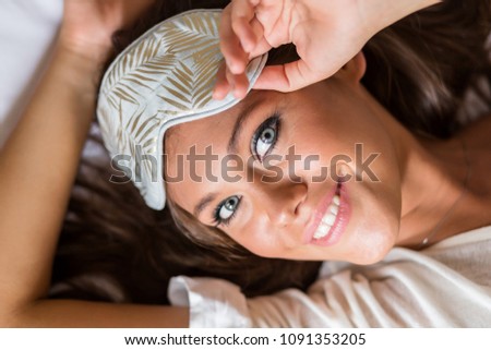 Young lady in pajamas and sleep eye mask with perfect skin posing at bedroom lying in bed after wake up and look to camera. Beautiful girl on white bed. Rest, sleeping, comfort and people concept