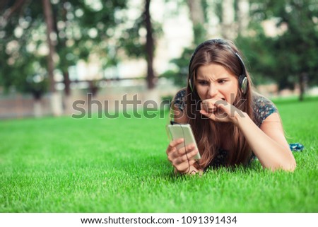 Young woman in a horror frustration bite his fist looking at phone screen outside in park lying down on green lawn background with copy space. Multicultural model mixed race asian russian teenage girl