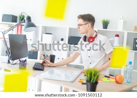 A young man with glasses and headphones stands near the table and works at the computer. A young man holds a marker in his hand and prints on the keyboard.