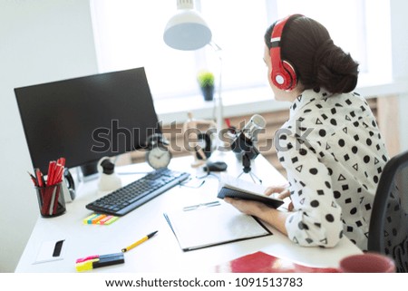 Beautiful young girl sits in headphones and with a notepad at the table in the office and speaks into the microphone.