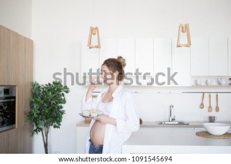 Pregnant woman in the kitchen holds a plate of cakes. A pregnant woman is standing in the kitchen in jeans and a white shirt Royalty-Free Stock Photo #1091545694