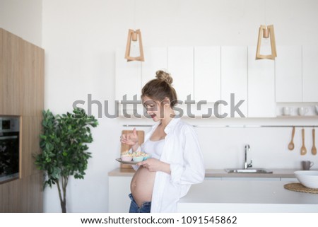 Pregnant woman in the kitchen holds a plate of cakes. A pregnant woman is standing in the kitchen in jeans and a white shirt. Royalty-Free Stock Photo #1091545862