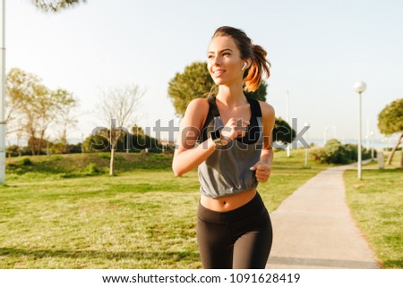 Photo of amazing young sports woman running outdoors on grass in park listening music with earphones.