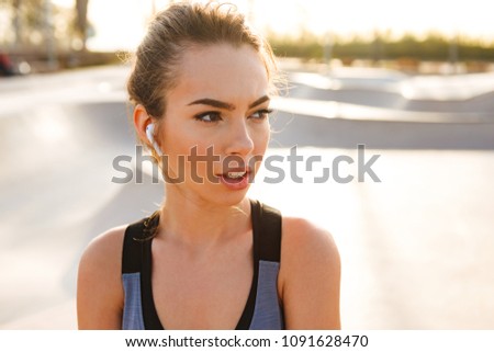 Picture of young sports woman standing outdoors listening music with earphones looking aside.