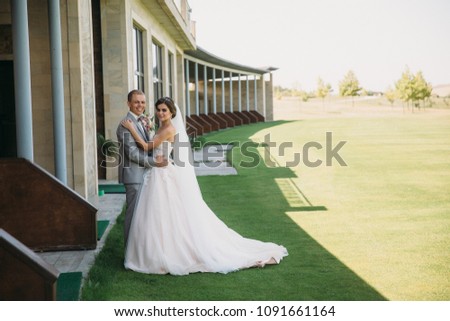 newlyweds are walking along the green field of the golf club on a wedding day. The groom in a business suit is gray and the bride in a luxury white dress with a veil are holding hands.