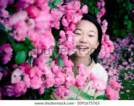 Outdoor portrait of beautiful young Chinese woman in yellow dress smiling among pink rose flowers wall in spring garden.