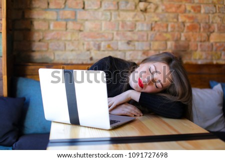 Bored female student with red lips sitting near modern laptop at cafe on sofa, brick wall background. Concept of learning information, using gadgets and free hotspot.