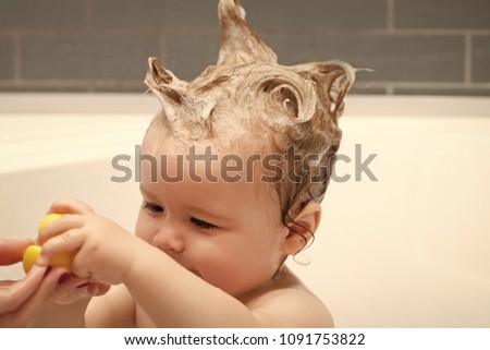 Little beautiful male kid sitting in white bathroom with wet foam hair taking yellow duckling toy from hand of mother, horizontal picture