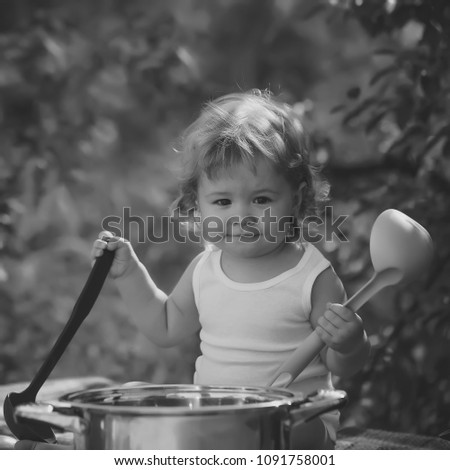Smiling boy with blonde curly hair in white underwear holding black and green kitchen utensil and cooking in big pot looking forward outdoor sunny day on natural background, square picture