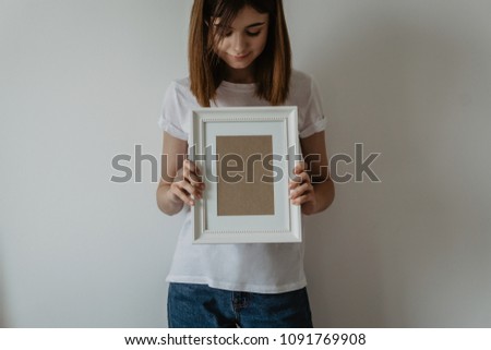 Young hipster girl in white t-shirt holding in hands empty vertical white wooden blank photo frame. Empty place for your photo, picture, gratitude letter, design or logo. Horizontal mockup.