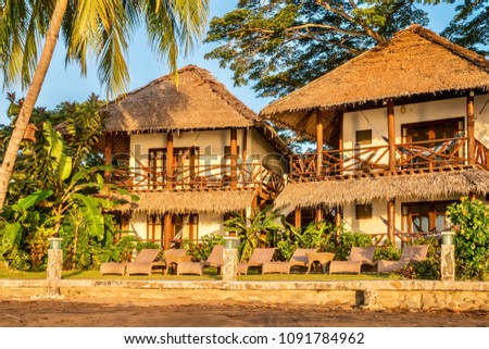 Beautiful beachfront resort in a tropical island beach setting, with rustic wooden features and traditional thatched roof design, surrounded by palm trees and greenery. Philippines.