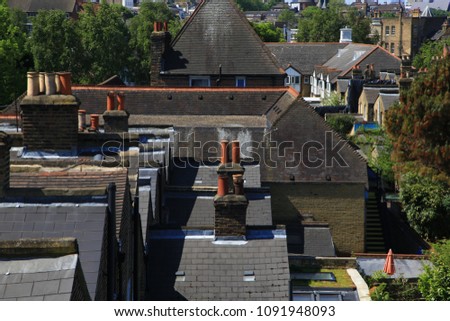 Rooftop view of residential housing in South London, England on a bright sunny day.  Sky is blue and trees are full and green amidst a sea of rooftops with chimneys.