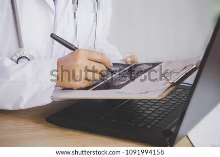doctor holding pen in hand analyzing x-ray medical picture while working on computer laptop on desk 