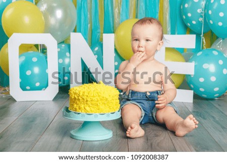 Portrait of cute adorable Caucasian baby boy in jeans pants celebrating his first birthday. Cake smash concept. Child kid sitting on floor in studio eating tasty yellow dessert 
