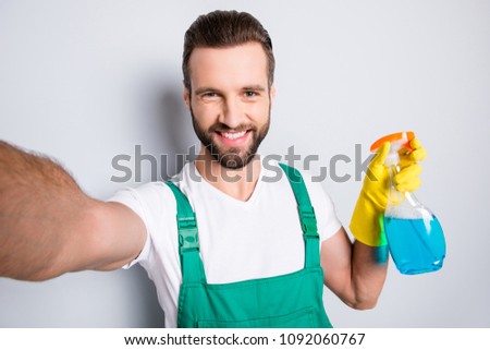 Self portrait of cheerful joyful cleaner with stubble in uniform shooting selfie on front camera having, showing detergent spray with blue liquid, isolated on grey background