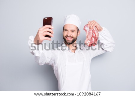 Portrait of attractive handsome butcher with stubble shooting selfie on front camera of smart phone, holding fresh meat in hand, blogger having video-call, isolated on grey background