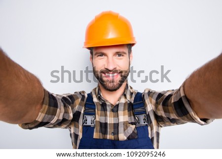 Portrait of handsome cheerful repairer in safety helmet with stubble shooting selfie on smart phone with two hands, wearing shirt and overalls, isolated on grey background