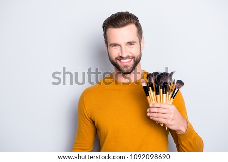 Portrait of handsome cheerful visagist with stubble in sweater having verity of brushes in arm, looking at camera, standing over grey background