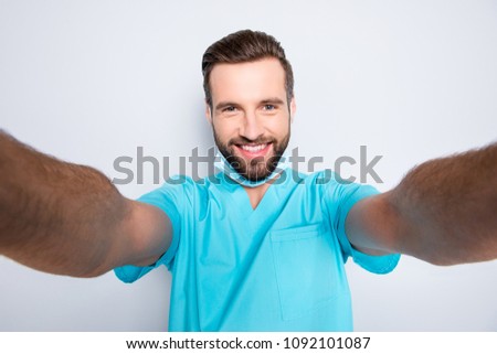 Self portrait of attractive cheerful man with stubble shooting selfie with two hands on front camera having sterile, protective, face mask, wearing lab blue uniform, isolated on grey background Royalty-Free Stock Photo #1092101087