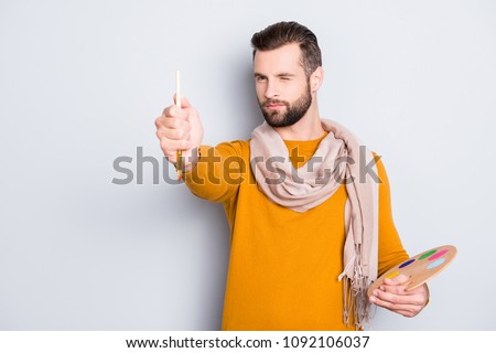 Portrait of creative concentrated artist with bristle in sweater and scarf on neck using, having colorful palette, brushed in arms, analyzing, expertising pic, isolated on grey background