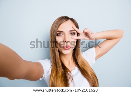 Self portrait of pretty cheerful girl shooting selfie on front camera gesturing v-sign near eye blowing kiss with pout lips isolated on grey background