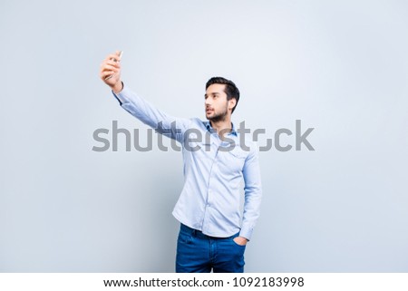 Portrait of trendy narcissistic stylish man holding hand in pocket of pants using smart cell phone shooting self picture on front camera isolated on grey background