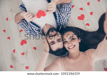 Self portrait of sweet cute couple lying head to head on the floor with small red hearts around shooting selfie on front camera, having fun leisure at home indoor