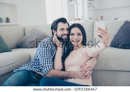 Portrait of lovely positive couple sitting on the floor indoor in house using smart phone shooting self portrait on front camera having video-call fun, enjoying time together