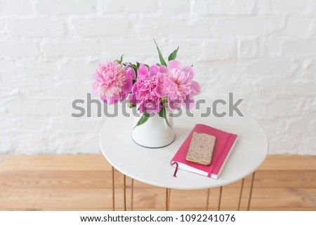 Three pink peony flowers in vintage jug on white coffee table on bright sunny day Notebook and mobile phone on the table top