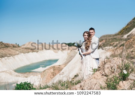 The bride in a beautiful dress hugging the groom in a light suit near the lake. Wedding couple standing on a sandy hill in the open air. A romantic love story. Azure-blue water on the horizon.