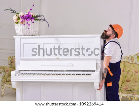 Loader moves piano instrument. Courier delivers furniture in case of move out, relocation. Delivery service concept. Man with beard, worker in overalls and helmet lifts up piano, white background