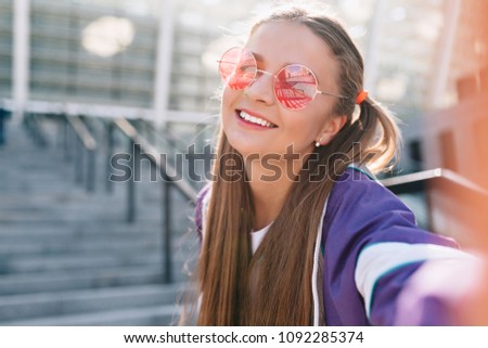 Wonderful charming girl in pink round glasses making selfie on sunny street on urban background. Photo of funny lady enjoying walking on summer street.