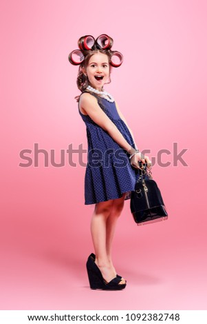 Portrait of a funny little girl with curlers in her hair in mother's shoes and bag. Studio shot over pink background. Kid's fashion. 