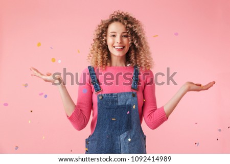 Portrait of a smiling young curly blonde girl playing with colorful confetti isolated over pink background