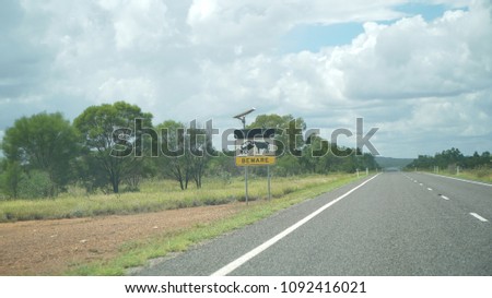 A sign stand on Flinders Highway, looks like ox is eating a car, careful about "car eating ox when lights on". Background with cloudy sky.