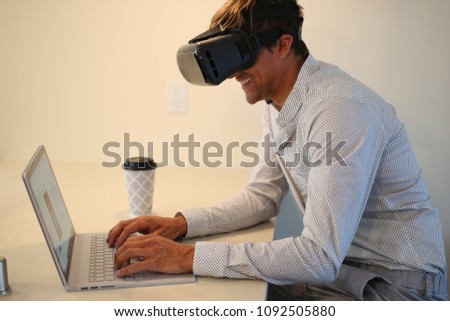 Young man wearing virtual reality glasses, with grey computer, coffee. Caucasian business looking, grey shirt.  Royalty-Free Stock Photo #1092505880
