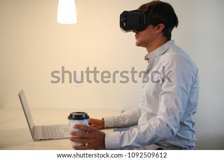 Businessman wearing virtual reality, augmented reality black glasses. Young man working on grey laptop, with cup of coffee. Inside the room, can be home or living room. Royalty-Free Stock Photo #1092509612