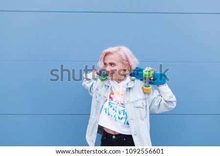 Happy girl stands with a skateboard against the background of a blue wall and smiles. Stylish blonde girl with a skateboard on a blue background.