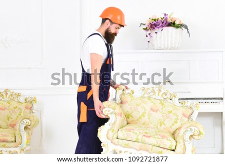 Relocating concept. Courier delivers furniture in case of move out, relocation. Loader moves armchair for move out. Man with beard, worker in overalls and helmet lifts up armchair, white background