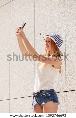 Technology, internet, social media concept. Young woman wearing sun hat taking picture of herself, selfie, with smartphone camera. Outdoor shot on sunny summer day