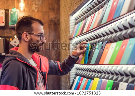 Choosing the right case for your smartphone. Young handsome man with glasses choosing case for his mobile phone in store Royalty-Free Stock Photo #1092751325