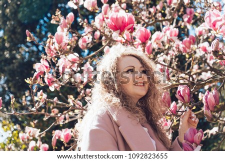 curly elegant young woman in a pink coat posing in a park in the sun next to a pink magnolia