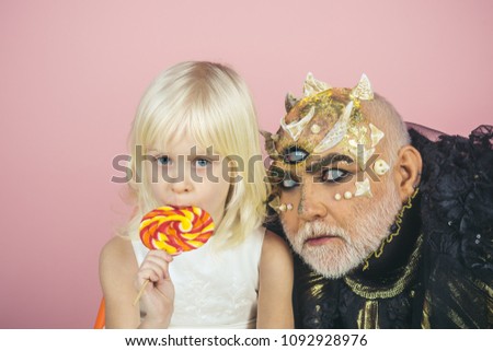 Innocent child with colorful lollipop embraced by old monster with thorns, warts and dragon skin. Lovely blond girl with male demon isolated on pink background. Fantasy world, good and evil concept.